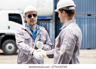 Two construction workers wearing hard hats and gloves shake hands in an industrial setting by a truck and shipping containers.