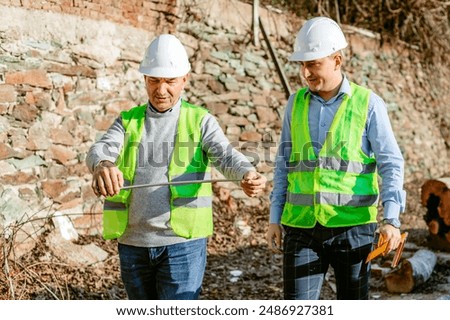 Two construction workers, wearing hard hats and safety vests, stand on a construction site. One worker holds a steel rod while the other observes. The workers appear to be measuring the length.