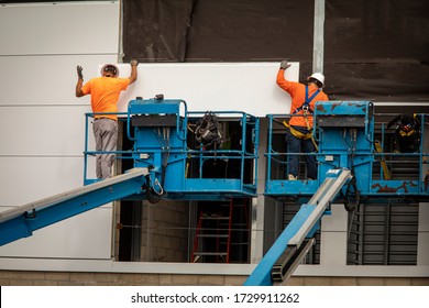 Two construction workers wearing hardhats on a blue manlift hanging section of drywall paneling on prefab building