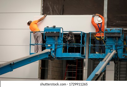 Two Construction Workers Wearing Hardhats On A Blue Manlift Hanging Section Of Drywall Paneling On Prefab Building