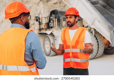 Two construction workers wearing orange safety vests and helmets at a quarry site near heavy dump truck. Teamwork concept