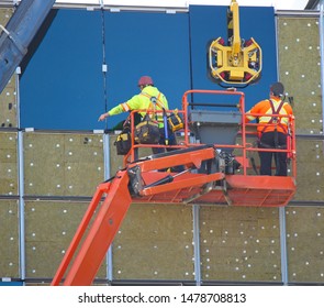 Two construction workers working at heights, safety harnessed to their sky lift.  One workman is signalling the crane operator to lower a large pane of glass. A machine holds the glass by suction.