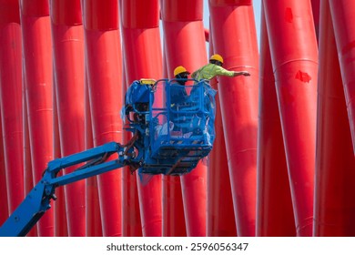 Two construction workers in yellow hard hats and green vests are lifted in a blue aerial work platform to work on the red steel structures of a bridge.