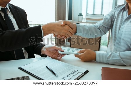 Two corporate businessmen shaking hands while one man places money on document in office room with corruption concept.