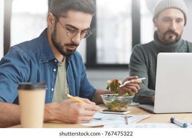 Two corporative workers work together at workplace: mature man keyboards on laptop and his companion writes attentively notes, eats fresh salad and drink coffee, have no time for going in cafe - Powered by Shutterstock