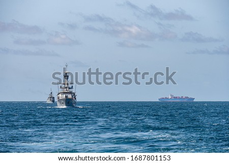 Two corvette warships sail together in the sea with container ship on the horizon.