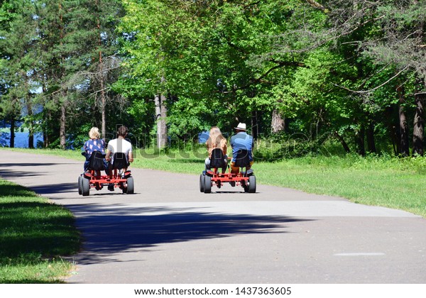 Two couples of adults\
women and men riding pedal kart car at park in sunny summer day,\
back view. Leisure time with friends and family, summertime\
vacation concept.\
