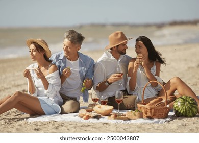 Two couples enjoying a sunny picnic on the beach, sharing food and wine, and engaging in cheerful conversation. Perfect moment of friendship and relaxation. - Powered by Shutterstock