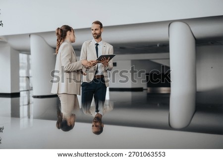 Two courteous professionals in light suits converse in a bright, modern lobby. A tablet is shared as they discuss plans, presenting a confident, collaborative business moment.