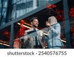 Two coworkers chatting on office balcony, sipping coffee and discussing business strategy with city view in background. Smiling and happy, they collaborate professionally