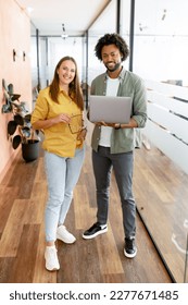 Two coworkers man and woman is using laptop, standing at the office. They holding laptop, looking at camera and smiling, full lengths, friendly office atmosphere and teamwork concept