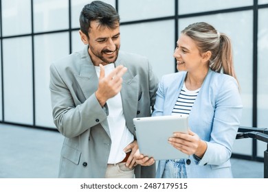 Two coworkers using a digital tablet and talking while standing outside. Modern business colleagues chatting together in front of office building on their way to work. Businesspeople collaboration - Powered by Shutterstock