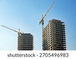 Two cranes are working on the construction of a tall building. The cranes are yellow and are positioned above the building. The sky is clear and blue, and the buildings are tall and imposing
