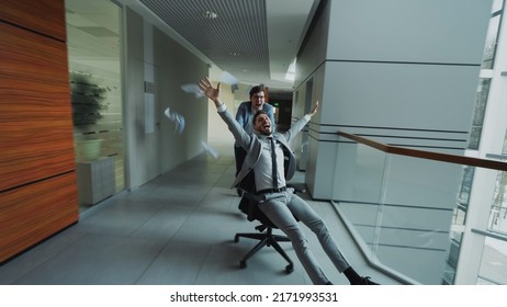 Two Crazy Businessmen Riding Office Chair And Throwing Papers Up While Having Fun In Lobby Of Modern Business Center Indoors