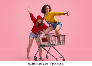 Two crazy female hipsters in bright outfits looking away while pushing and riding trolley against pink background - Shutterstock ID 1442095643