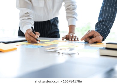 Two Creative Business People Meeting And Planning Use Post It Notes Sticky Note On Desk To Share Idea, Analysis Data Chart And Graph With Teamwork Strategy Brainstorming In Office.