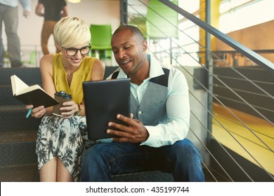 Two creative millenial small business owners working on social media strategy using a digital tablet while sitting in staircase
