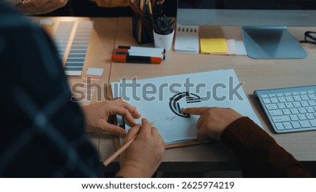 Two creative professionals discuss a logo design, surrounded by color palettes and stationery in a modern office space, showcasing collaboration and artistic inspiration. SACTR