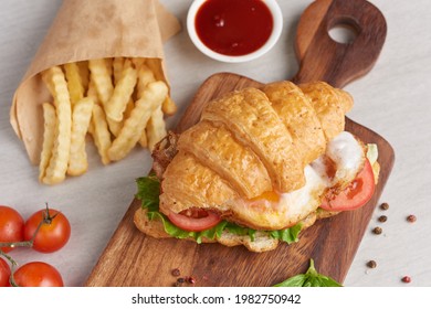 Two Croissant Sandwiches On Wooden Table, Top View, Sandwich With Bacon, Fried Egg. Ham, Cheese, Bacon, Fried Egg, Tomato, French Fries And Lettuce Served On Wooden Cutting Board.