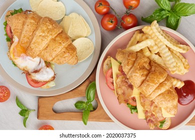 Two Croissant Sandwiches On Wooden Table, Top View, Sandwich With Bacon, Fried Egg. Ham, Cheese, Bacon, Fried Egg, Tomato, French Fries And Lettuce Served In A Plate.