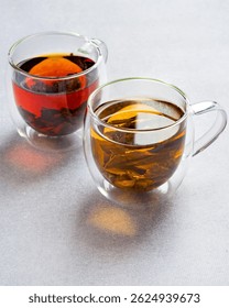 Two cups of assorted black and green tea in transparent double glassware on light grey background.