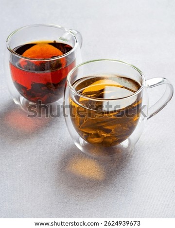 Two cups of assorted black and green tea in transparent double glassware on light grey background.