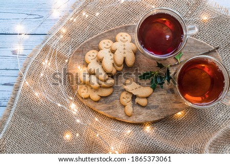 two cups of black tea and a stack of gingerbread cookies or ginger-man on brown canvas with Christmas lights on a white table 