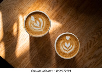 Two cups of fresh coffee with latte art on wooden table in sunlight. Morning cappuccino with hearts in cafe for couple. Coffee break concept. Caffeine drink for breakfast closeup. Top view, flat lay.  - Powered by Shutterstock