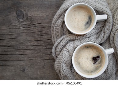 Two Cups Of Hot Coffee With Foam In The Shape Of A Heart On A Rustic Wood Table Background Wrapped In Warm Knit Wool Scarf. View From The Top. Space For Text. The Concept Of Heat. Holidays And Events.