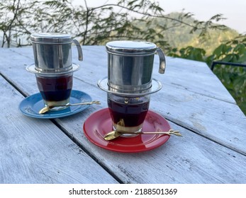 Two Cups Of Indonesian Coffee, With A Stainless Filter Above The Glass. Drinks On A Wooden Table Against A Backdrop Of Natural Scenery And Trees. Black Coffee Mixed With Milk.