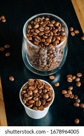Two cups with roasted coffee beans - one transparent glass and one small paper cup on the black and brown wooden table in the cafe