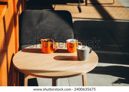Two cups and a teapot on a table in a cafe with hard shadows. Orange teapot and cups in the interior of a modern cafe. Sunny morning and cups of tea. Copy space.