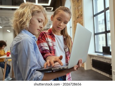 Two curious kids, little boy and girl using laptop while standing in a classroom during STEM lesson
