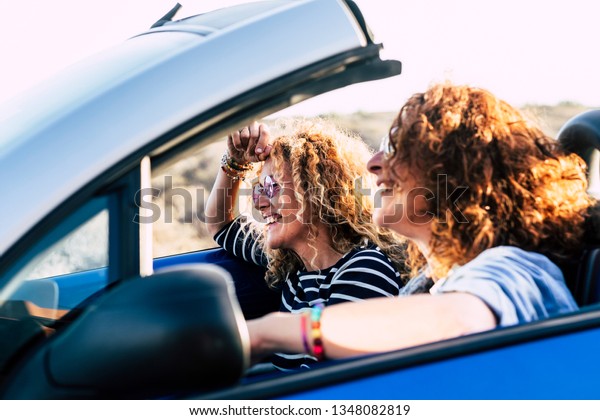 Two curly beautiful women friends drive and travel\
together on a convertible blue car having fun - outdoor happy\
leisure activity for cheerful people under the sun of summer -\
focus on second girl