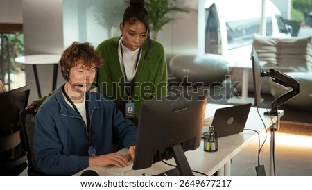 A two customer support professionals are working together modern office environment. one is seated, wearing headset, and typing computer, while other stands nearby, attentively observing screen