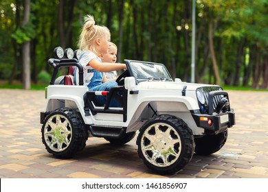 Two Cute Adorable Blond Sibings Children Having Fun Riding Electric Toy Suv Car In City Park. Brother And Sister Enjoy Playing And Driving Vehicle On City Street Outdoor. Happy Childhood Concept