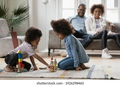 Two Cute African American Kids Playing Wooden Blocks On Floor In Living Room, Building Toy Constructions, Practicing Learning Games At Home. Black Couple Of Parents Resting On Couch, Watching Children