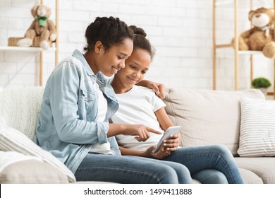 Two cute african american sisters using phone on sofa at home, older sister pointing at smartphone screen, copy space