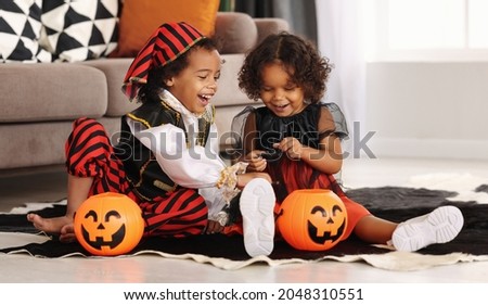 Two cute african kids brother and sister in Halloween costumes playing with artificial spider toy while sitting on floor with orange pumpkin lantern during All Saints Day Party at home