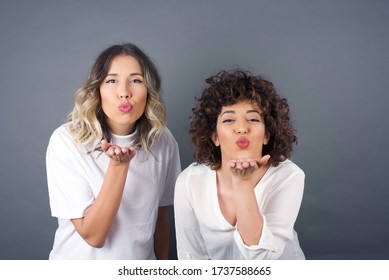 Two Cute Appealing Girl Friends Blow Kiss At Camera,  Or Says Goodbye On Distance, Isolated Over White Studio Background. Attractive Young Women Shows Sympathy To Someone.