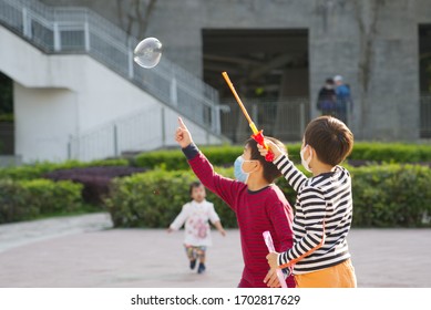 Two Cute Asian Boy With Disposal Surgical Mask Is Playing The Bubble Making Toy At The Playground With Fun Under Sunshine.Kid Free Play,outdoor Exercise And Learning During COCID-19/corona Concept.