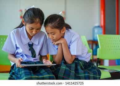 Two cute Asian elementary schoolgirls, long hair in a white shirt dress, skirt and green plaid tie are playing games on the tablet easily in school.