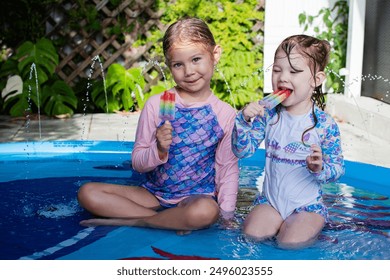 Two cute Caucasian children eating rainbow popsicle in a pool. Kids summer fun concept. Happy childhood during summer break. Kids in a splash pad eating ice cream - Powered by Shutterstock