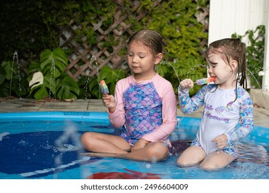 Two cute Caucasian children eating rainbow popsicle in a pool. Kids summer fun concept. Happy childhood during summer break. Kids in a splash pad eating ice cream - Powered by Shutterstock