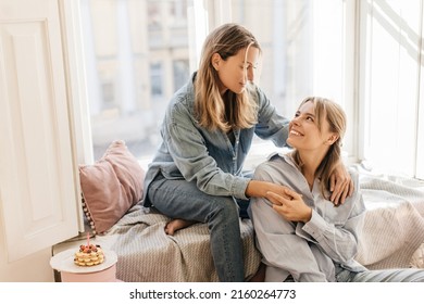 Two Cute Caucasian Mother And Daughter Look At Each Other Sitting At Window During Day. Blondes Wearing Casual Clothes Spend Time In Family Circle. Concept Of Love And Care