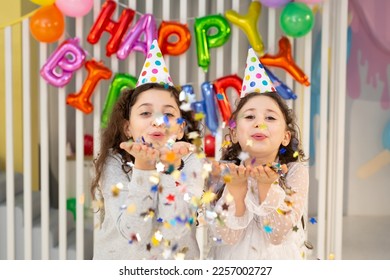 Two cute girls blow confetti on the background of a bright wall with balloons at a children's birthday party - Shutterstock ID 2257002727