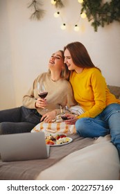 Two cute happy funny women friends having fun together, arranging home cinema evening, watching comedy on laptop, laughing out loudly, sitting on bed with cheese plate and glasses of red wine