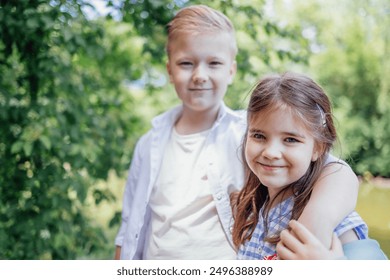 Two cute kids in casual clothes hug and pose for camera. Brother and sister smile and have good time together in nature. Little girl and boy against background of green trees. Vacations concept. - Powered by Shutterstock