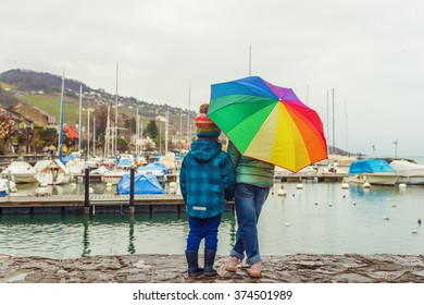 Two Cute Kids Resting By The Lake On A Rainy Day, Hiding Under Big Colorful Umbrella, Back View