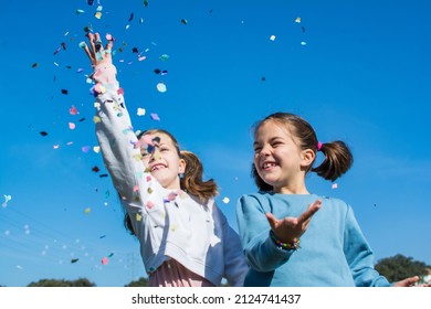 Two Cute Little Girls Celebrating And Playing With Confetti. Girls With Confetti And Streamers, One Is Blonde And The Other Is Brunette And They Have Pigtails. They Are In A Field With A Blue Sky.